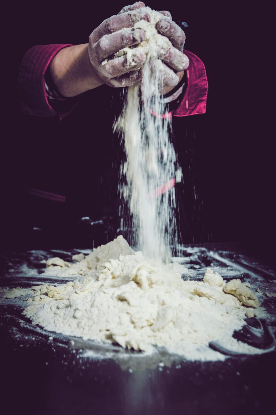 Two flour-covered hands releasing a stream of flour onto a heaped pile on a dark bench. Photo by Jordane Mathieu on Unsplash.