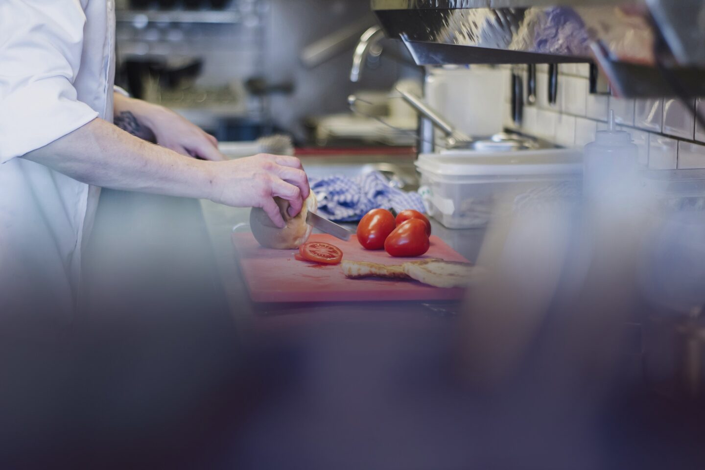 A chef in whites slicing tomatoes on a red board at a stainless steel bench, the kitchen soft behind them. Photo by Louis Hansel on Unsplash.